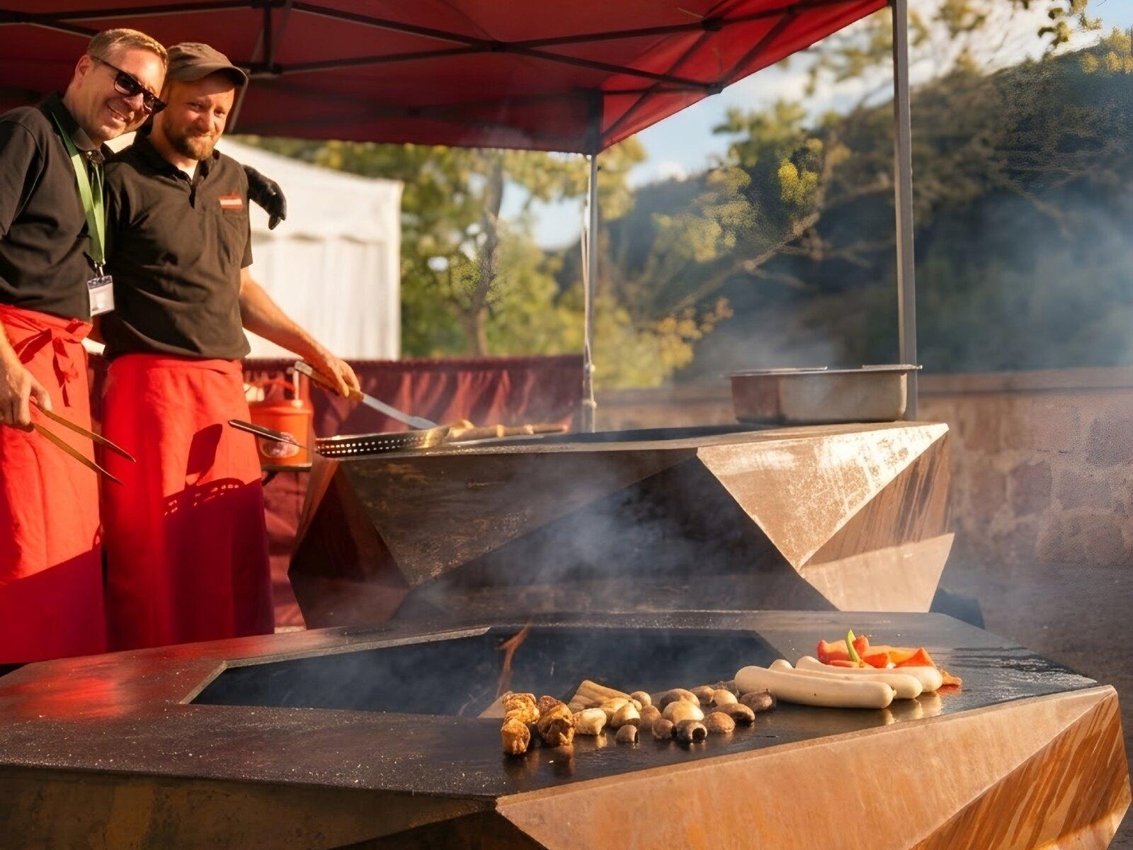 Two cooks in red aprons under a gazebo at two fire bowls, sausages and mushrooms on the plate in front