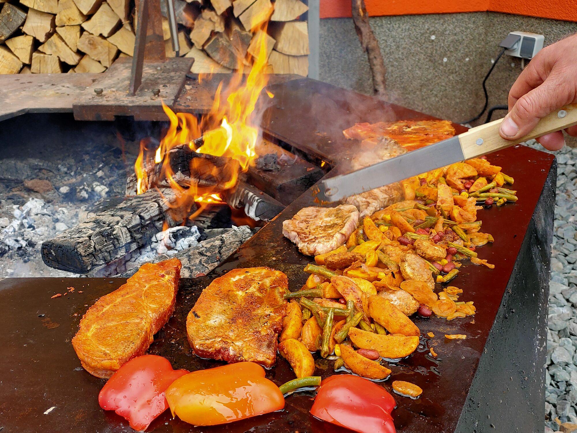 Steaks, peppers and vegetables cooking on the wide steel rim of the fire bowl beside the open embers